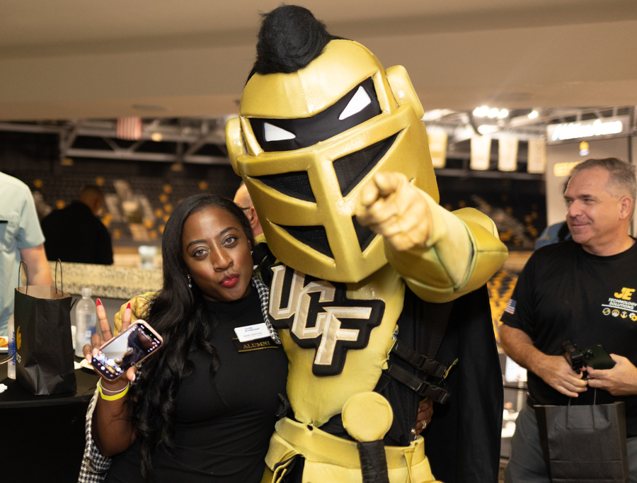 Woman posing with a person in a gold knight mascot costume featuring UCF insignia at an indoor event, holding a smartphone.