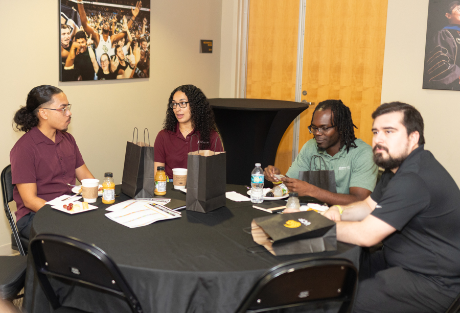 Four people sit around a round table with a black tablecloth, enjoying snacks and drinks while bags and papers are on the table in a casual gathering.