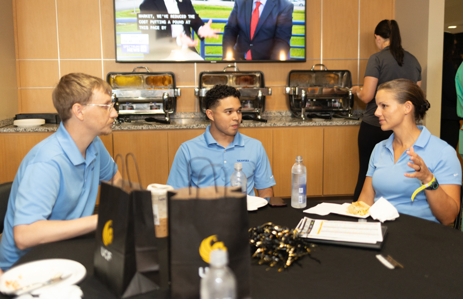 Three coworkers in blue polo shirts sit at a table with takeout bags and water bottles, conversing in a buffet area behind them.