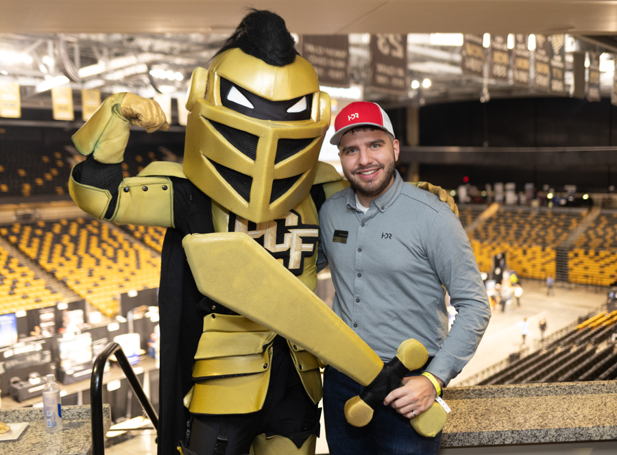 Man in a gray polo and red cap posing with a large gold knight mascot inside a sports arena with yellow seats in the background