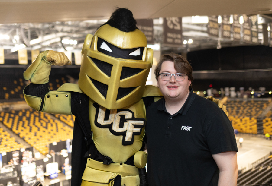 Person and gold UCF Knights mascot pose together in an arena with yellow seating in the background.
