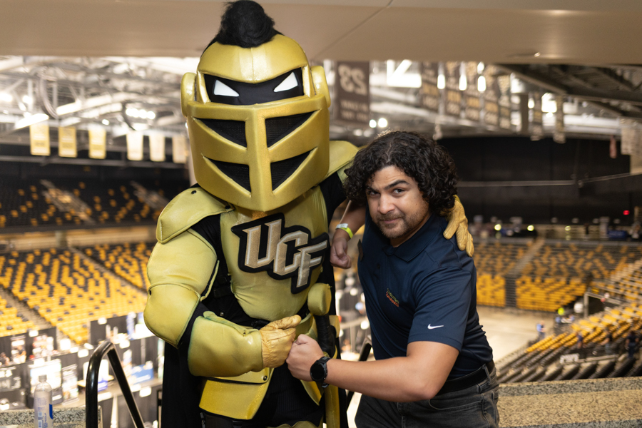 Man posing with a gold UCf Knights mascot in a large indoor arena filled with yellow seats.
