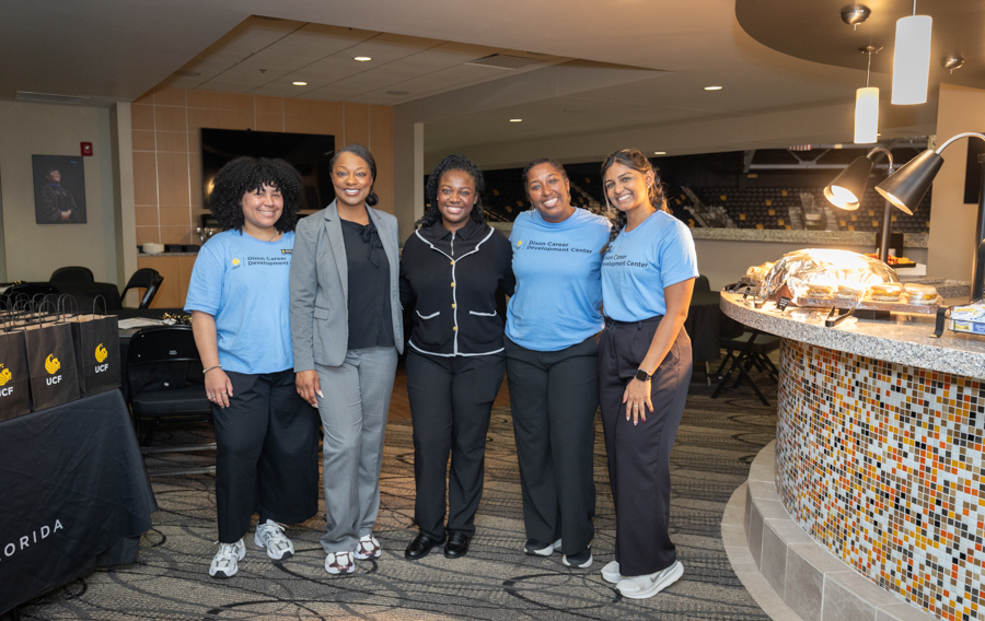 Five women posing together in a modern lounge, with a curved mosaic food counter visible to the right and some in blue staff shirts in the group to the left a few in business attire.