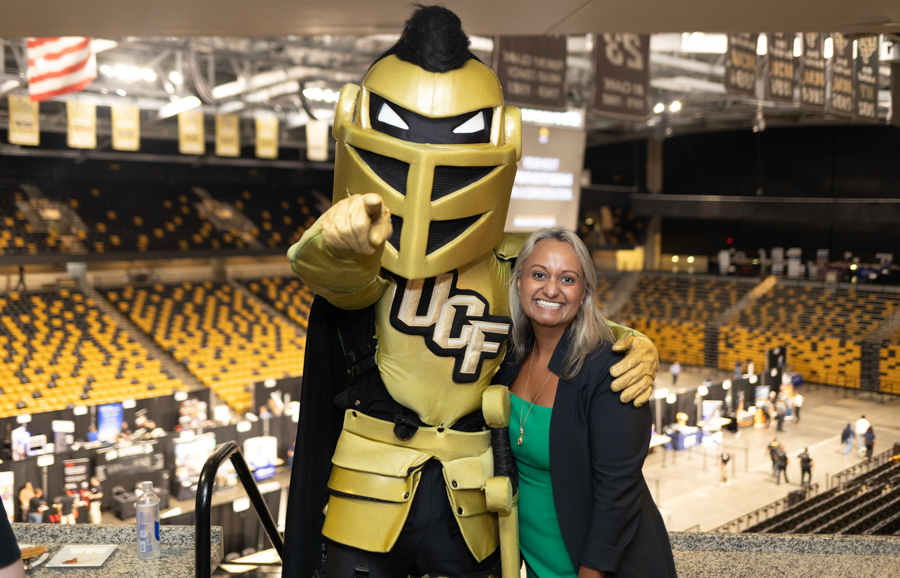 UCF Knight mascot in gold armor poses with a smiling woman in a green top inside a large indoor arena with yellow and black seats.