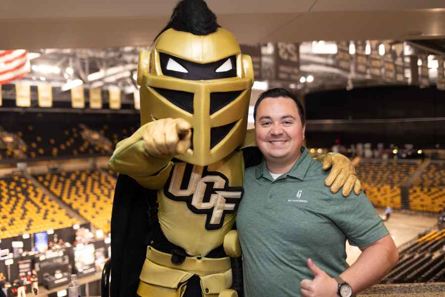 UCF Knight mascot in gold armor posing with a smiling man in a green polo inside a stadium arena.