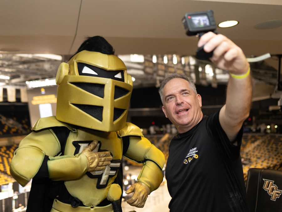 Man in a black T-shirt takes a selfie with a large gold knight mascot inside a sports arena.