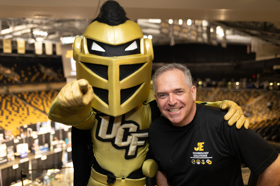 Man smiles beside a gold UCf knight mascot in an arena, pointing at the camera with his arm around the mascot.