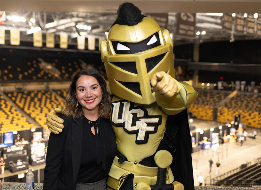 Smiling woman posing with a gold UCF Knight mascot in a large indoor arena, both looking at the camera.
