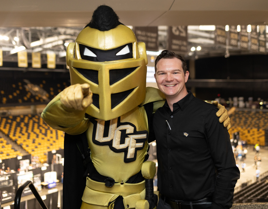 Smiling man in a black shirt posing with a gold UCF Knights knight mascot inside a sports arena.