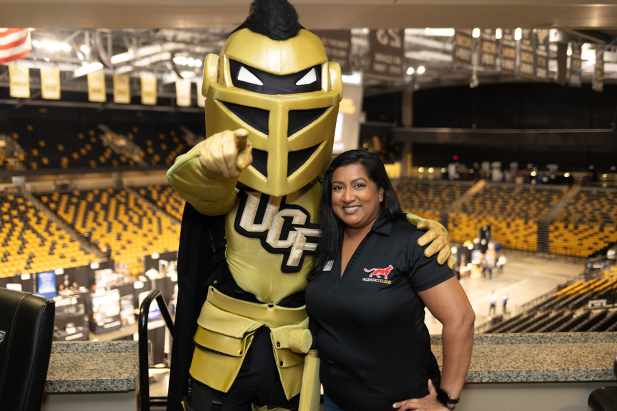 UCF Knights mascot in gold armor poses with a smiling woman in a black polo inside a large indoor arena at a game or event.