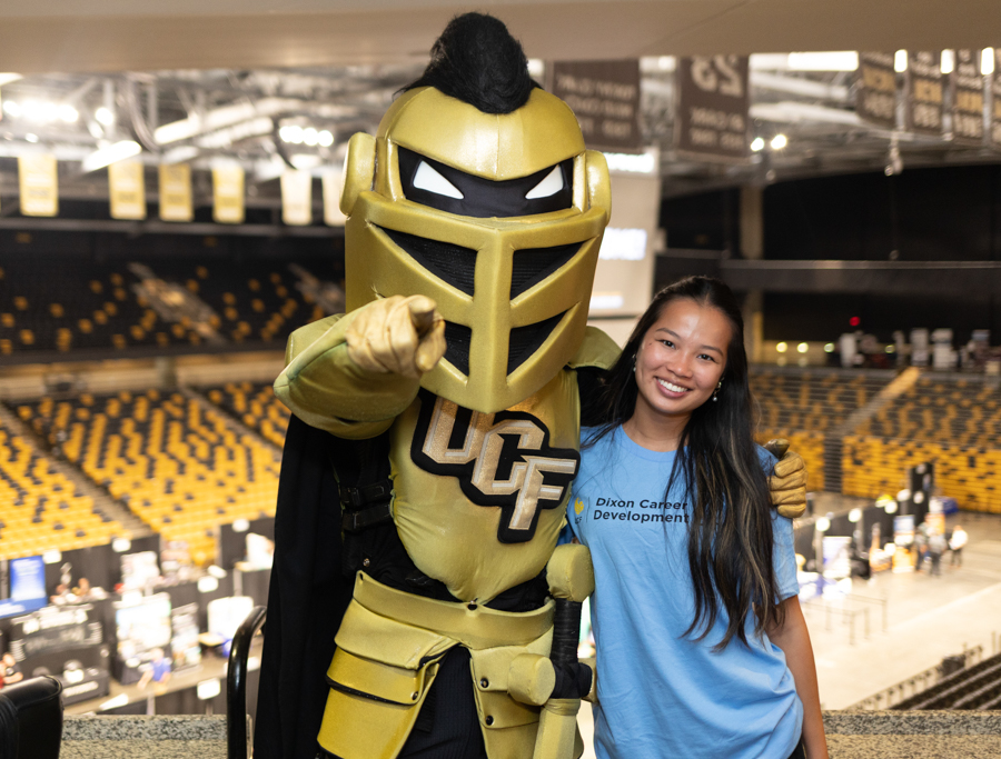 UCF mascot in gold armor points at the camera beside a smiling woman in a blue shirt at a stadium with yellow seats in the background, cheering event scene.