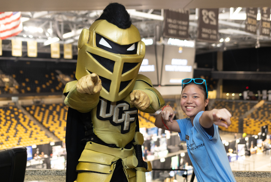 UCF Knights mascot in gold armor points at the camera beside a smiling student in a blue shirt in a stadium concourse. - or - Mascot in gold armor and a student in a blue shirt strike a playful pointing pose in an arena.