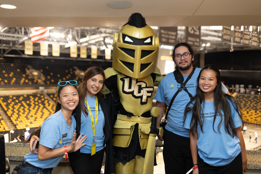 Group of four students posing with a gold UC mascot in a large sports arena.