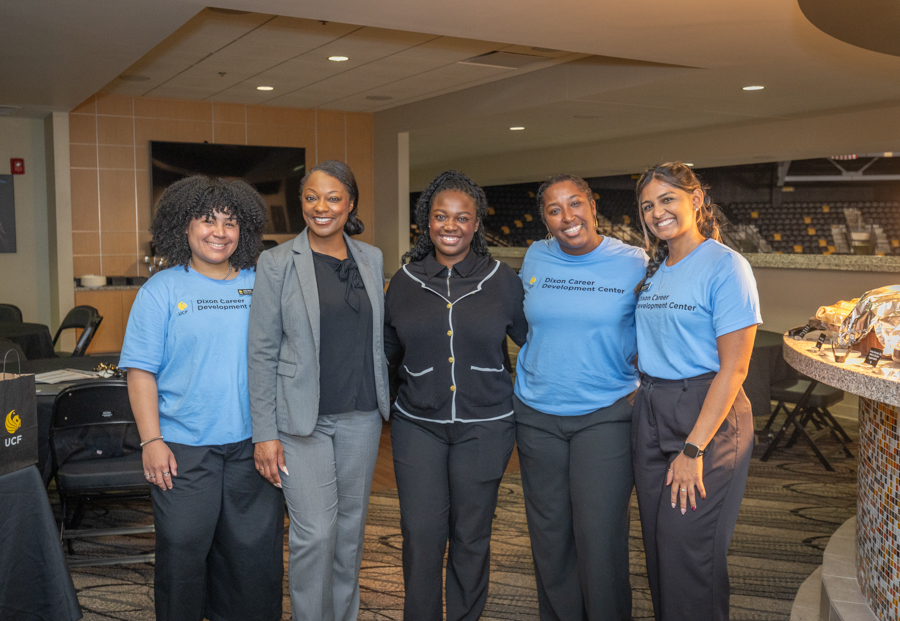 Five women posing together indoors for a group photo at an event; two wear blue Dixon Career Development Center shirts and others in business casual attire, all smiling.