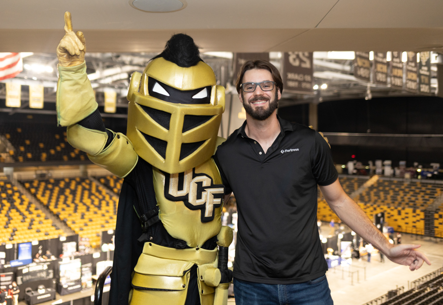 Man in a black polo and safety glasses posing with a gold knight mascot in a stadium. A gold armored knight mascot with raised finger beside a man in black polo inside an arena. Smiling man stands next to a gold UCF knight mascot in a large indoor arena. Two people posing for a photo: knight mascot and man, with yellow seating behind.