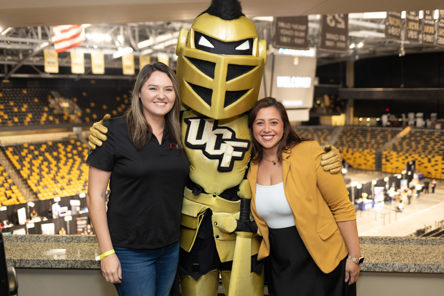 Two smiling women pose with a life-size gold UCF Knights mascot in a sports arena.