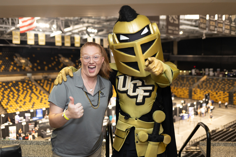 Smiling woman in a gray polo gives a thumbs up beside a gold knight mascot in an arena with yellow seats in the background.