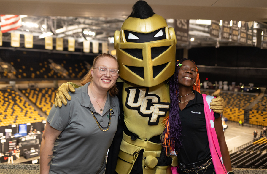 UCF Knights mascot with two smiling fans posing in a large indoor arena with yellow seats behind them.