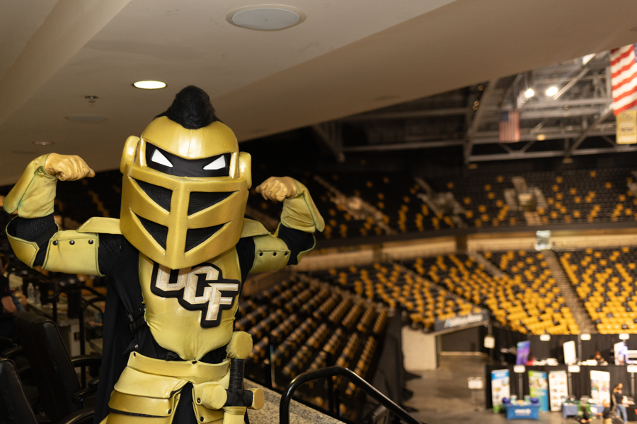 Golden knight mascot in gold armor flexing arms in a large arena with yellow and black spectator seats.