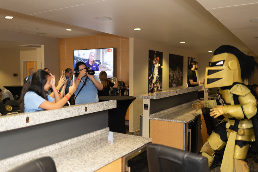 Costumed knight in gold armor greets fans at a lobby/bar area while people take photos with cameras and phones nearby.