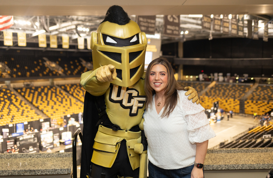 UCF Knights mascot in gold armor posing with a smiling woman in a white blouse inside a sports arena with yellow seats nearby.