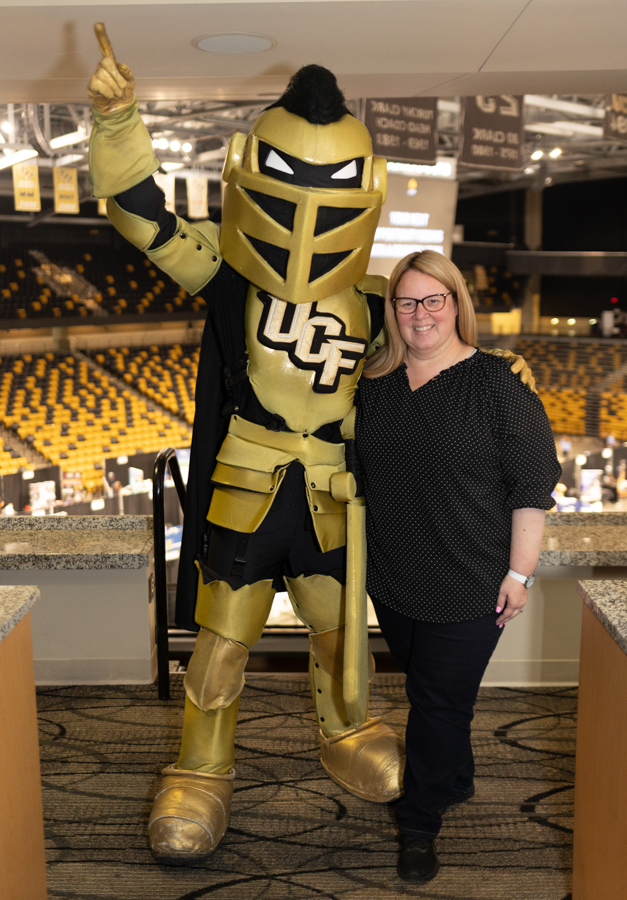 Smiling woman posing with a large gold UCF Knight mascot in a sports arena with yellow seating behind them.
