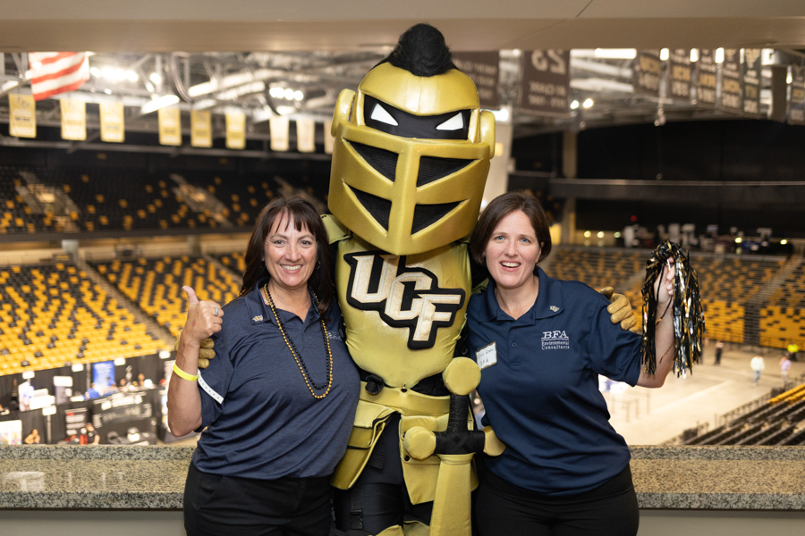 Two smiling women posing with a large gold knight mascot in a sports arena with yellow seating and banners in the background, UCF logo on the costume.
