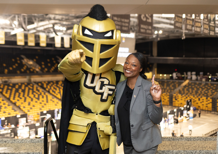 UCF Knight mascot in gold armor posing with a smiling woman in a gray blazer inside an indoor arena.