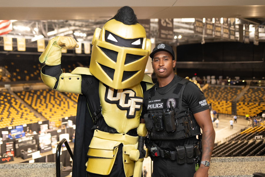UCF Knights mascot in gold armor posing with a police officer in a large arena with yellow seats.
