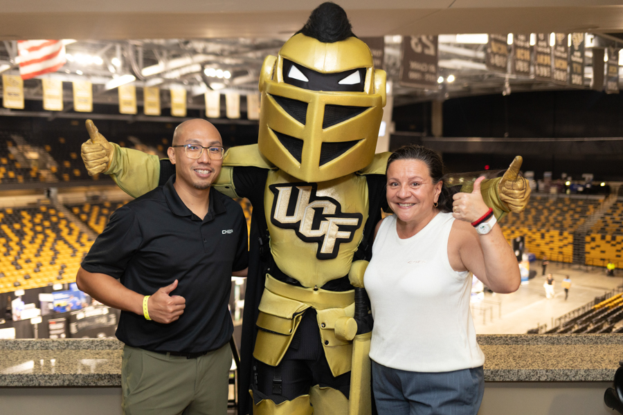 Man and woman posing with a large UCF Knight mascot in a sports arena, both giving thumbs up and smiling.