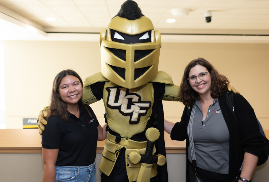 Two women pose with a large gold knight mascot wearing armor and a UCF logo in a campus lobby, smiling at the camera.