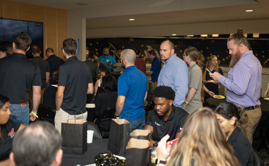 People gathered at a busy indoor event, standing around tables with gift bags and coffee cups in a conference hall.