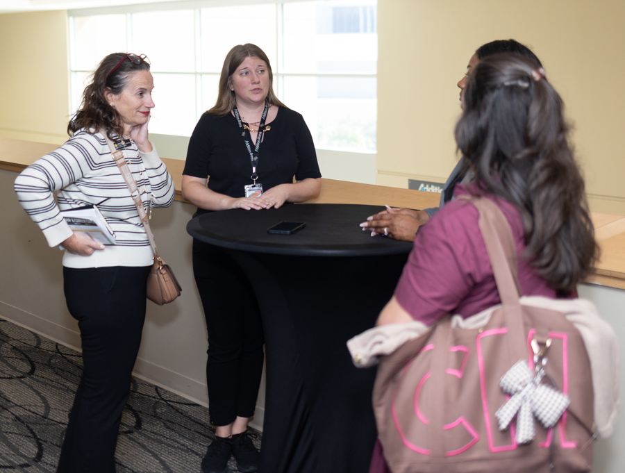 Four people stand around a tall round table in a bright hallway, engaged in discussion; one woman holds papers and wears a striped sweater while another speaks with a lanyard around her neck.
