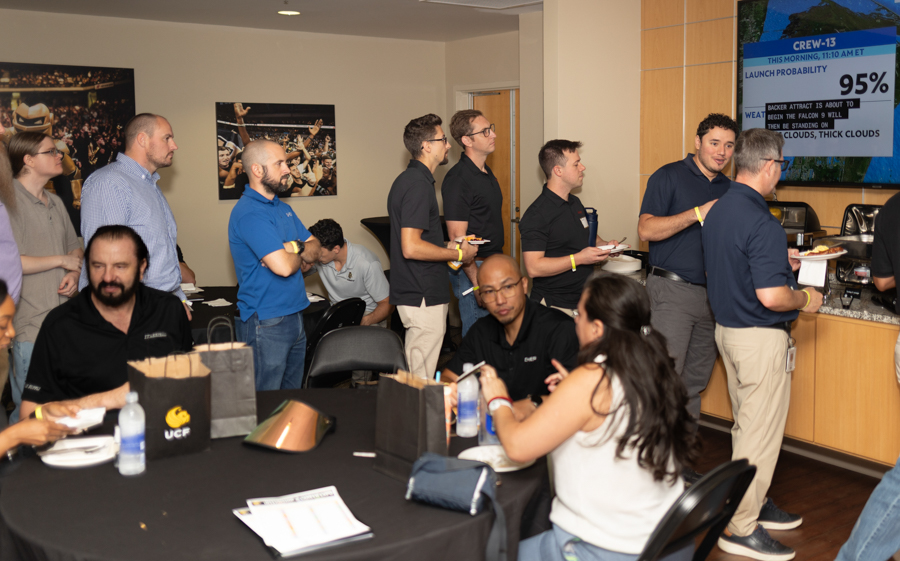 Office social event: coworkers line up for food at a buffet while others sit at a round table, a presentation screen visible in the background.