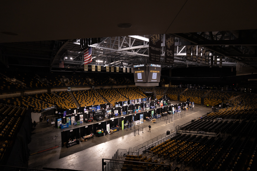 Wide view of a large arena transformed into an expo floor with numerous booths along a central row of tables and attendees milling about.