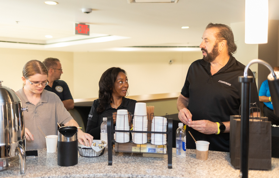 Barista with a beard talks to two customers at a coffee counter with cups and utensils nearby.