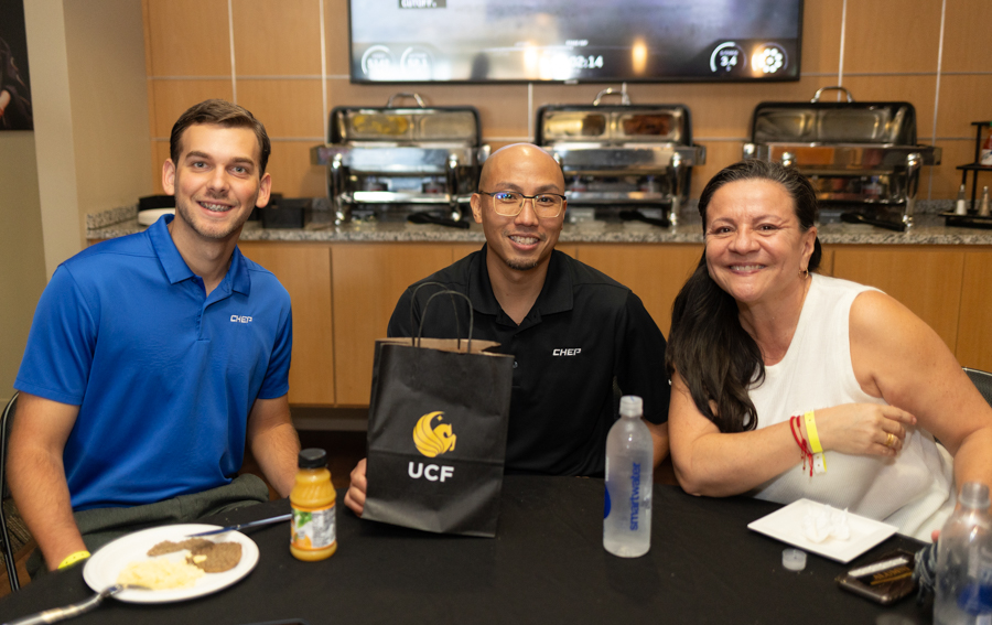 Three people sit at a catered event table and smile at the camera, with chafing dishes and a counter behind them.