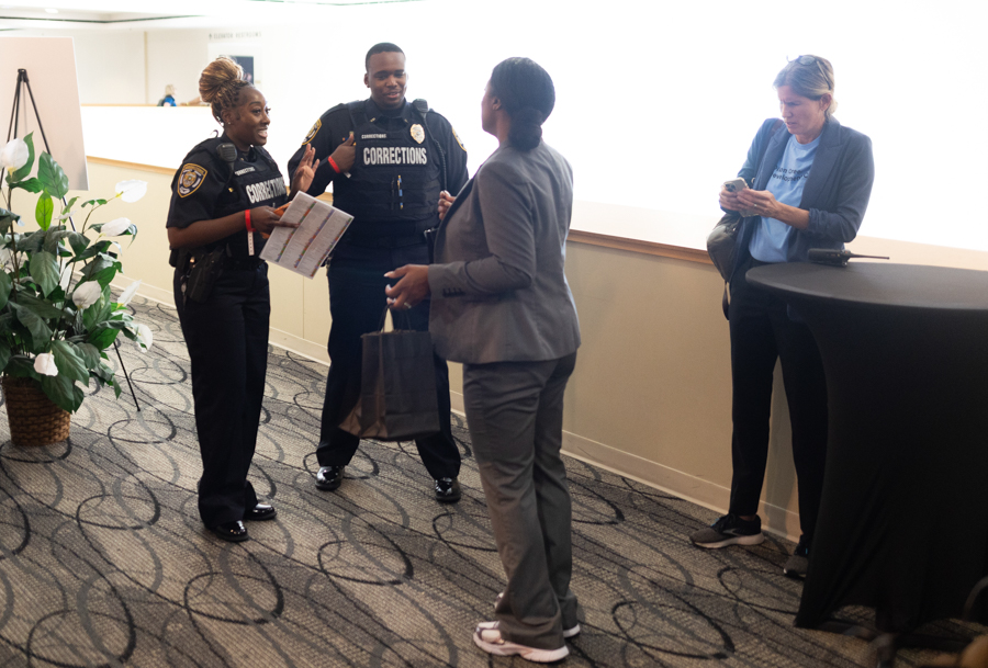 Two corrections officers speak with a woman in a gray suit who carries a shopping bag in a hallway near a tall table.