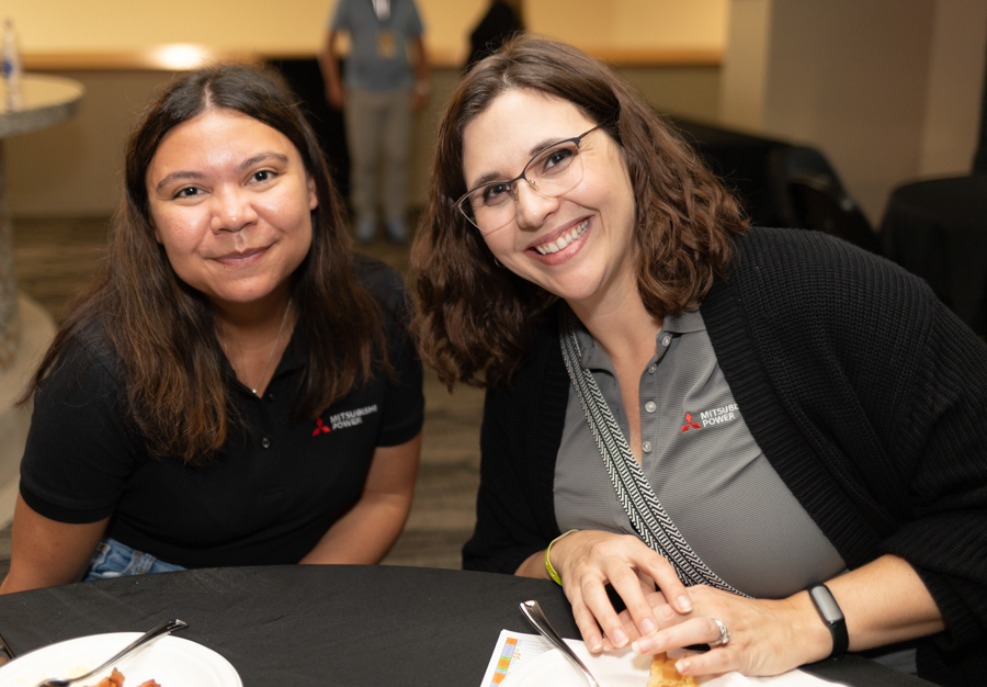 Two women smiling at a table, posing for a photo at an event; one wears glasses and a gray Mitsubishi Power polo while the other wears a black polo.