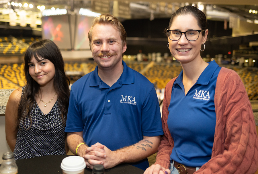 Three people smile at the camera in a large arena, two in blue MKA polo shirts and a third in a patterned top, with a coffee cup on the table in front.