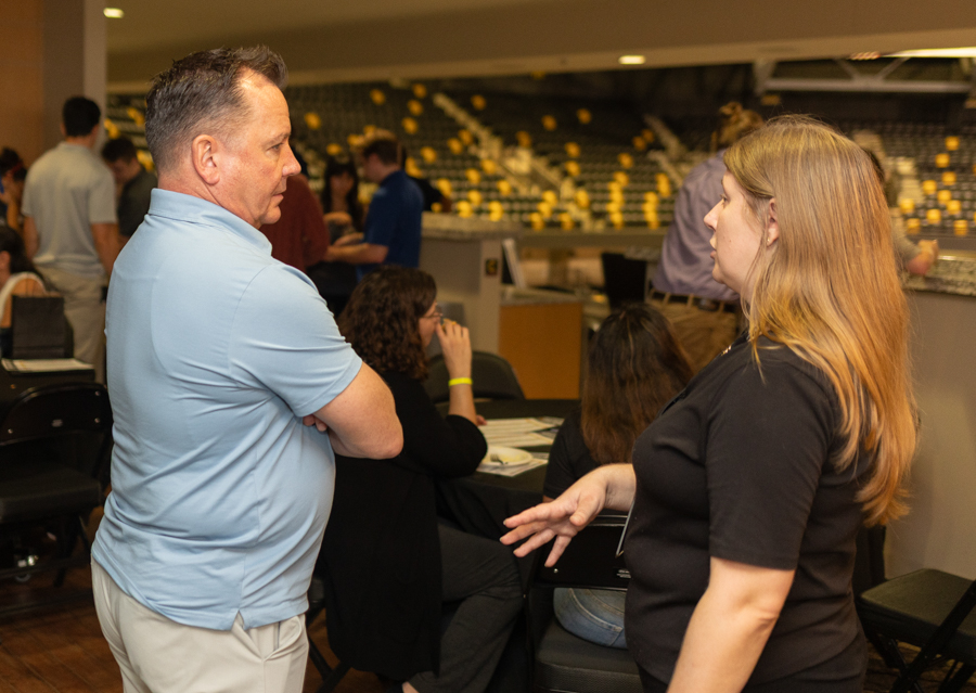 Man and woman conversing at a social/event gathering in a large hall, with other attendees seated at tables in the background.