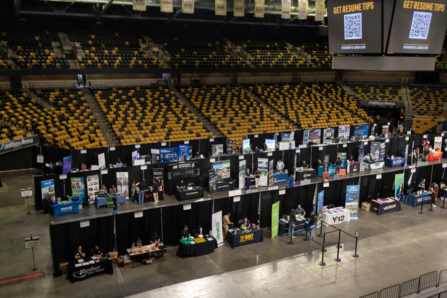 Wide shot of a career fair inside a large arena with rows of booth tables and yellow seating in the background.
