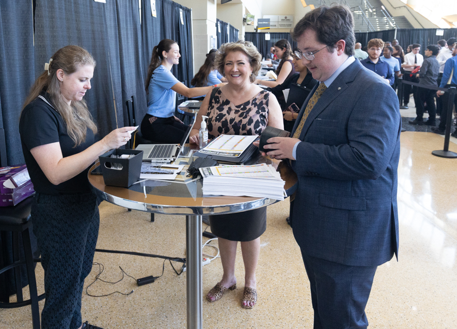 Registration desk at a career fair; a smiling woman in a leopard-print top hands materials to a man in a suit as he signs in/fills forms nearby.