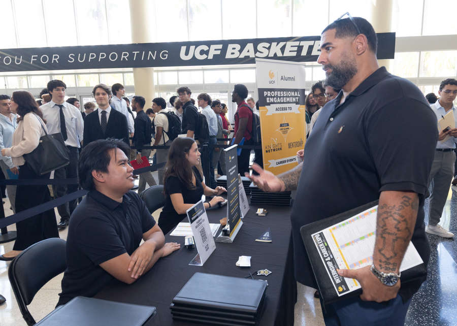 Recruiters sit behind a registration table at a UCF career fair, talking to a visitor with a clipboard.