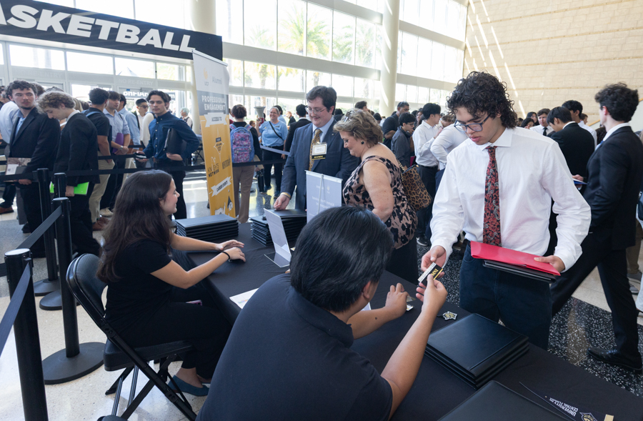 Registration desk at a busy conference or career fair with attendees and staff handing out badges and folders at a long table in a bright atrium.