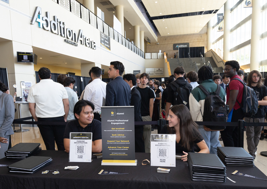Registration desk at a campus career fair in a bright atrium, with students queueing and two volunteers seated behind folders and signs.