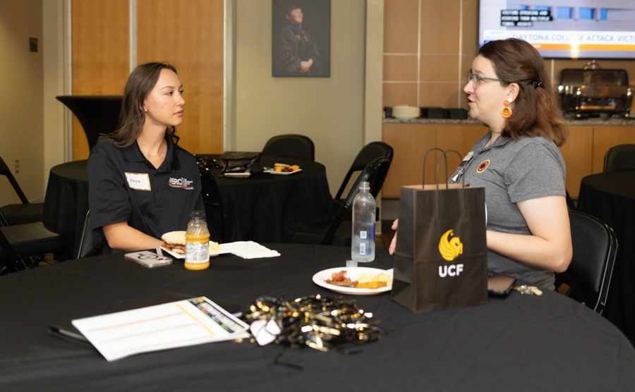 Two women sit at a round table in a conference room, having a conversation, with a UCF-branded bag, water bottle, and snacks on the table.