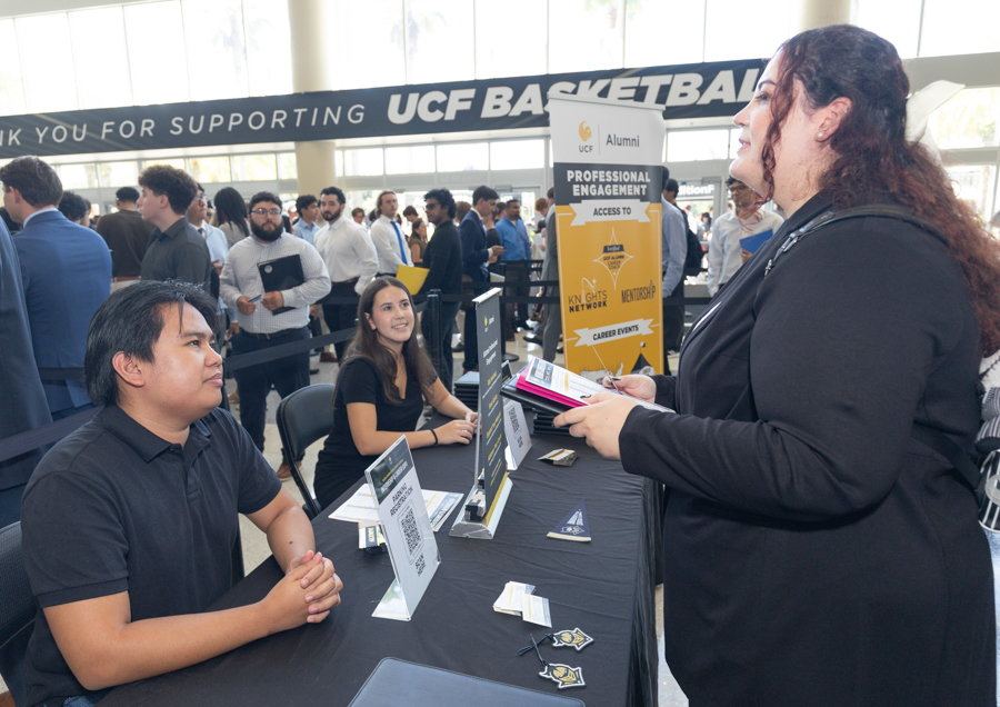 Recruiter at a university career fair greets a job seeker at a table with signs and flyers.