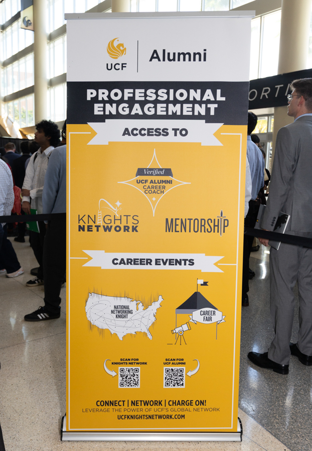 Tall yellow conference banner for UCF Alumni highlighting professional engagement, access to mentorship and career events in a bustling lobby.
