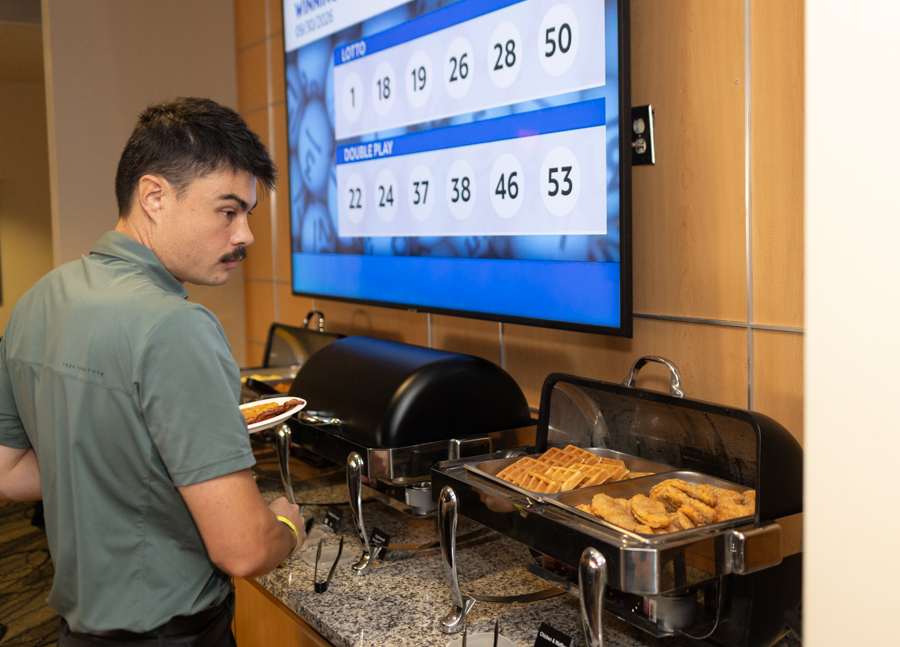 Man at a breakfast buffet with waffles and fried chicken, looking at a large screen displaying lottery numbers in the background.
